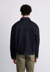 Giubbotto in denim nero con colletto classico, cuciture visibili e bottoni in metallo. Il retro presenta un taglio dritto e un'etichetta patch.