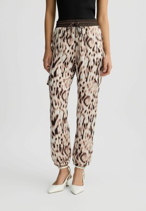 Kobieta w spodniach cargo w panterkę ze ściągaczem w talii i białymi szpiczastymi szpilkami, stojąca na jednolitym tle.