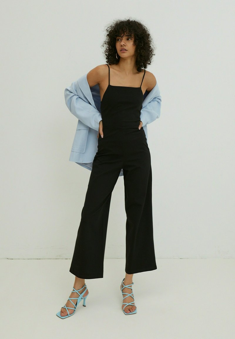 Zwarte jumpsuit met dunne bandjes, brede benen, gecombineerd met een lichtblauwe oversized blazer en blauwe hakken met bandjes.