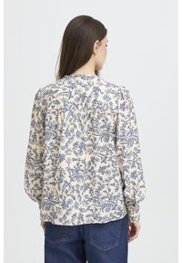 Blouse à manches longues florale en crème avec des imprimés botaniques bleus, dotée d'un dos boutonné, d'un design froncé et d'une texture de tissu douce.