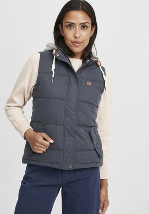 Gilet rembourré marine avec capuche, doté de cordons beige et de boutons-pression. Porté sur un pull léger, avec des poches avant et une coupe décontractée.