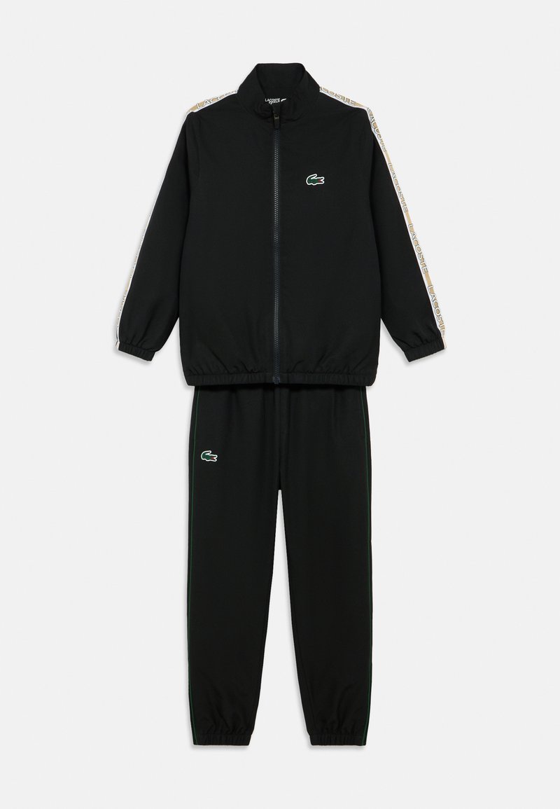 Crni trenirka od glatkog materijala. Jakna na patent s visokim ovratnikom, logotipom na prsima i uzorkovanom bočnom prugom. Hlače s elastičnim pojasom u istom uzorku.