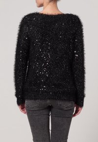 Svart fluffig tröja med paljetter, lös passform, vid halsringning, långa ärmar, texturerad yta; bärs med grå jeans.