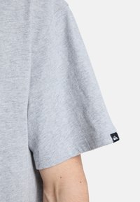 Γκρι κοντομάνικο t-shirt με απαλή υφή, με μια μικρή μαύρη ετικέτα λογότυπου στο αριστερό μανίκι. Απλός, καθημερινός σχεδιασμός.