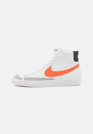 Hvid højtopsneaker med grå tåhætte, lysende orange Nike-swoosh, hvide snørebånd og sort hælflig med hvid tekst.