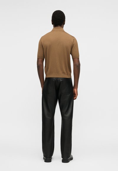 Άνδρας που στέκεται με την πλάτη, φοράει ένα καφέ κοντομάνικο πουκάμισο και μαύρο δερμάτινο παντελόνι σε απλό φόντο.