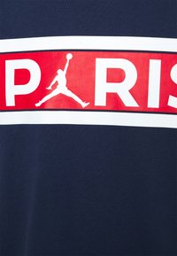 Námořnická modrá tričko s výraznou červenou a bílou obdélníkovou grafikou, na které je nápis "PARIS" a silueta basketbalového hráče. Hladká textura.