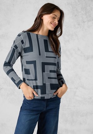 Camicia a maniche lunghe in tessuto blu navy con motivi geometrici bianchi, dotata di un collo rotondo e una vestibilità rilassata. Abbinata a jeans blu scuro.
