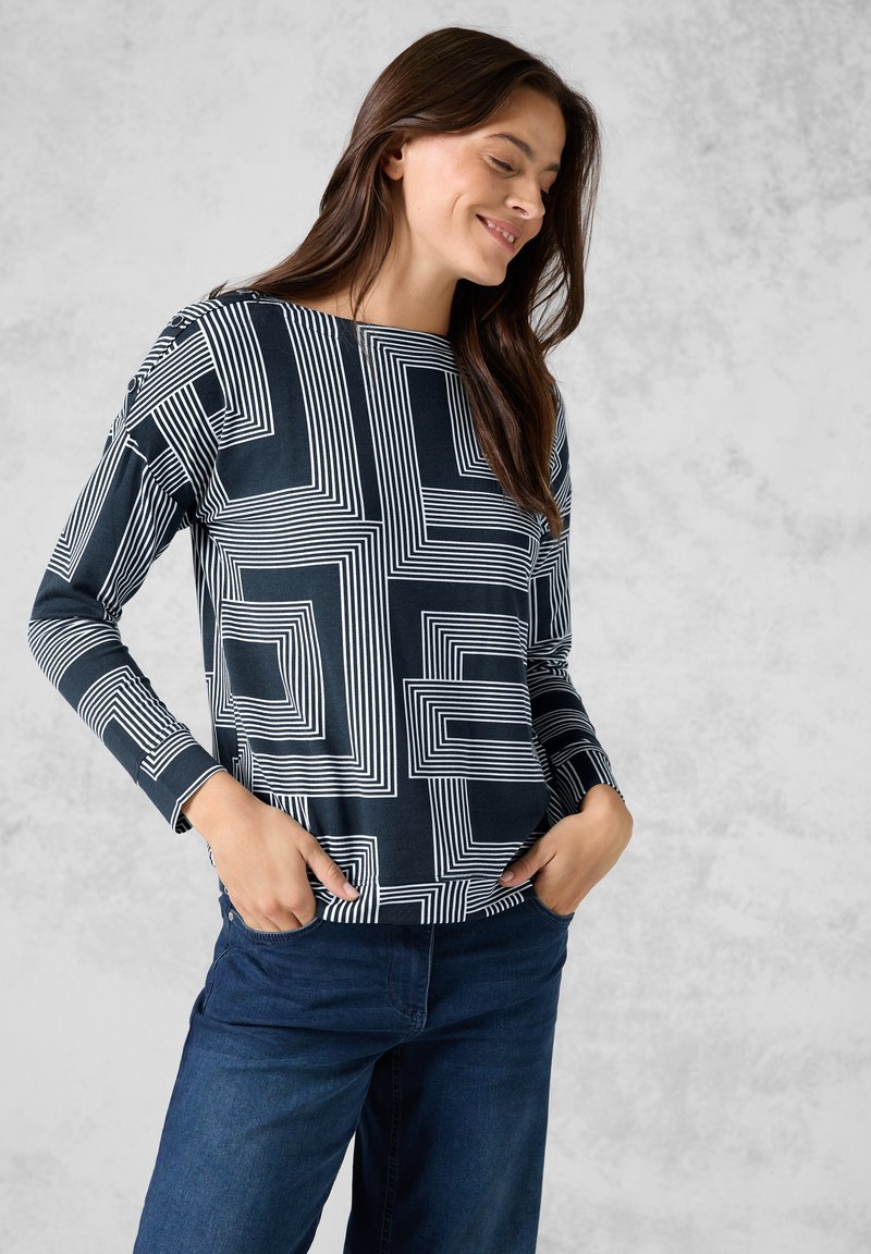 Camicia a maniche lunghe in tessuto blu navy con motivi geometrici bianchi, dotata di un collo rotondo e una vestibilità rilassata. Abbinata a jeans blu scuro.