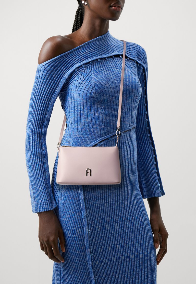 Borsa a tracolla rosa chiaro con texture liscia e logo metallico, indossata sopra un abito blu a costine con design off-shoulder e dettagli di bottoni.