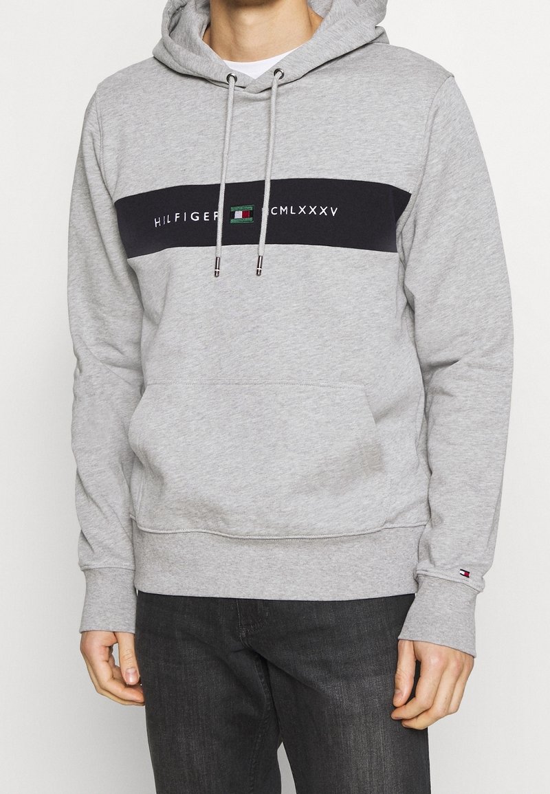 Homme portant un sweat à capuche gris clair avec une bande noire horizontale et le texte logo "HILFIGER CMLXXXV" sur la poitrine, associé à un pantalon foncé.