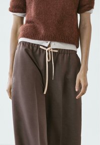 Hnědé kalhoty s širokými nohavicemi a měkkou texturou, se stahovací šňůrkou v světlejší barvě. Spárováno s vínově červeným chlupatým svetrem.