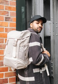 Beige hátizsák felső fedéllel és állítható pántokkal, tartós anyagból készült. Oldalsó zsebe és sima textúrája van.