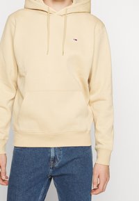 Sudadera beige con bolsillo frontal, capucha con cordón y pequeño logo. La tela parece suave; los puños y el dobladillo son acanalados. Se lleva con vaqueros azules.
