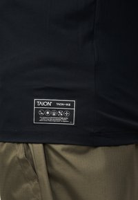 Μαύρο στρίφωμα πουκάμισου με λευκή ετικέτα της μάρκας TAION που δείχνει εικονίδια φροντίδας και κείμενο πάνω από χακί παντελόνι.