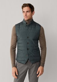 Dunkelgrüne, gesteppte Weste mit Stehkragen, Knopfleiste vorne und zwei Seitentaschen, getragen über einem hellbraunen Langarmshirt.