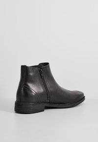 Botín de cuero negro hasta el tobillo con cremallera lateral, punta redondeada, tacón bajo y textura suave, con acentos mínimos y un diseño elegante.
