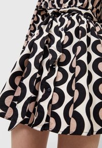 Kort klänning med en plisserad design har ett djärvt mönster av svarta och beige cirklar. Tyg är lätt och klänningen har ett knytmidja och knappdetaljer.