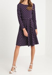 Kvinna klädd i en lila, långärmad klänning i knälängd med vita prickar och ett tygbälte, kombinerat med vita öppna klacksandaler.