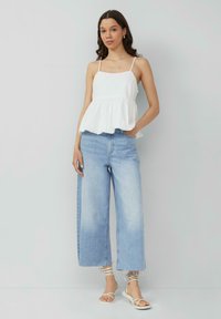 Biały top bez rękawów w fasonie peplum z regulowanymi ramiączkami; zestawiony z jasnoniebieskimi spodniami typu wide-leg i białymi sandałami na paski. Gładka tekstura materiału.