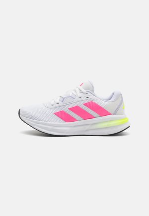 Zapatilla deportiva blanca con franjas rosas, parte superior de malla ligera, suela acolchada y una suela exterior de goma negra con un acento amarillo brillante.