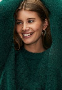 Zöld kötött pulóver, amelynek texturált, bolyhos felülete van. Kerek nyakkivágással készült, és ezüst karika fülbevalóval van párosítva.