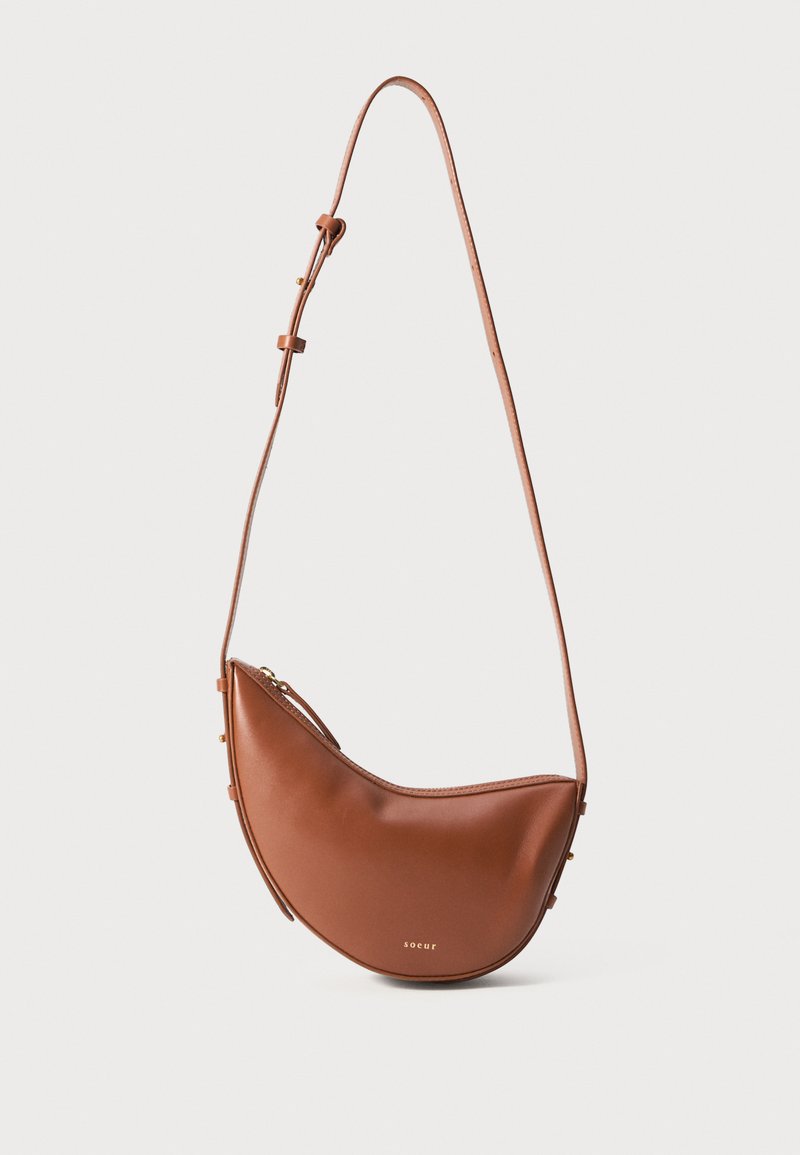 Bolsa de ombro em couro castanho, em forma de meia-lua, com alça ajustável e pequeno logótipo dourado "soeur" na parte da frente.