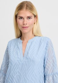 Bluză de culoare albastru deschis cu decolteu în V și un model texturat în valuri; are mâneci lungi transparente și o croială lejeră, realizată dintr-un material ușor.