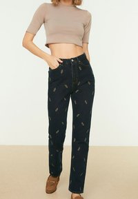 Vaqueros oscuros con bordado de hojas doradas, diseño de cintura alta, ajuste recto, combinados con una blusa corta de color marrón claro y zapatos marrones.
