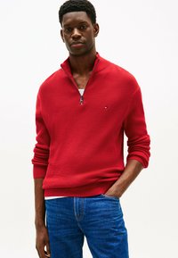Pull en tricot rouge avec un col zippé, en matériau texturé et à manches longues. Porté sur une chemise blanche, associé à un jean en denim bleu.