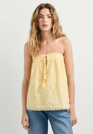 Giovane donna con lunghi capelli mossi, indossa un top smanicato ricamato giallo pallido con spalline sottili e jeans blu su uno sfondo semplice.