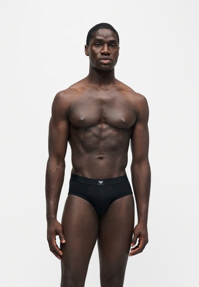 Homme musclé debout portant un sous-vêtement noir avec un petit logo sur la taille, sur un fond clair uni.