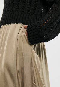 Hand in der Tasche eines beigen Plisseerocks, kombiniert mit einem schwarzen Strickpullover mit Durchbrüchen an den Ärmeln.