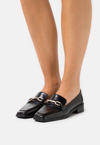 Svarta läderloafers med fyrkantig tå, som har en metallisk detalj över ovandelen och en låg blockklack för stabilitet.