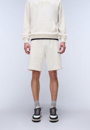 Mann trägt einen cremefarbenen Hoodie und passende Shorts mit schwarzen und weißen Sneakers und steht vor einem einfarbigen hellen Hintergrund.