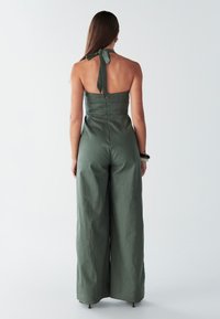 Groene linnen jumpsuit met een halternek, detail met strik aan de achterkant en een wijde pijp. Gestructureerde stof met een gladde afwerking.