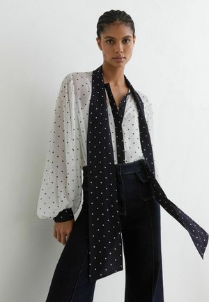 Donna con capelli intrecciati che indossa una camicetta bianca con pois neri, una lunga sciarpa nera abbinata e pantaloni scuri a vita alta.