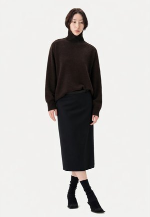 Femme portant un pull à col roulé marron foncé, une jupe crayon midi noire, des chaussettes noires et des chaussures noires à bouts pointus, debout sur un fond blanc.
