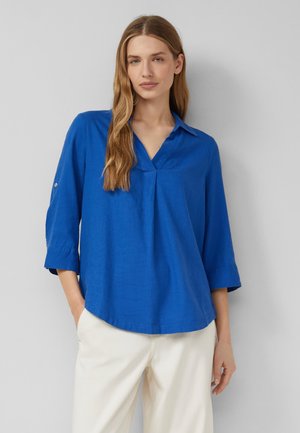 Kobieta o długich blond włosach, ubrana w luźną niebieską bluzkę z rękawami 3/4 i białe spodnie, stojąca na jasnoszarym, jednolitym tle.