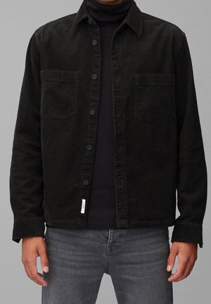Homme portant une veste en velours côtelé noire sur un col roulé noir, associée à un jean gris, debout devant un fond gris clair uni.
