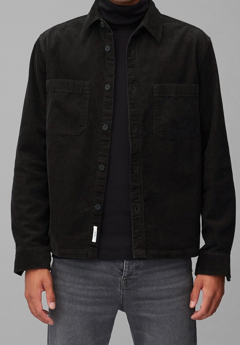 Homme portant une veste en velours côtelé noire sur un col roulé noir, associée à un jean gris, debout devant un fond gris clair uni.