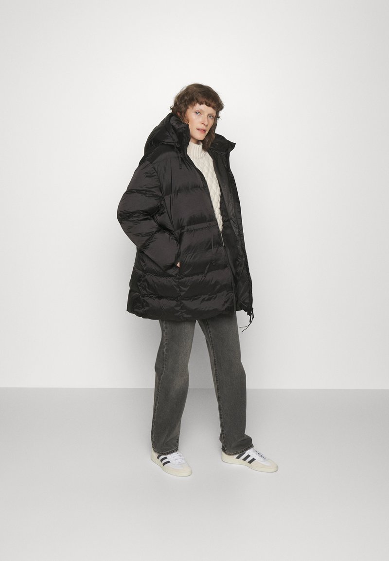 Schwarze, voluminöse Jacke mit hohem Kragen und Reißverschluss, kombiniert mit einem cremefarbigen, kabelgestrickten Pullover und grauen Hosen, abgerundet mit weißen Sneakers.