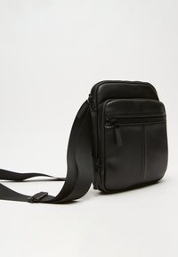 Borsa a tracolla nera in pelle con due scomparti con zip, tracolla regolabile e superficie texturizzata. Forma rettangolare con accenti minimi.