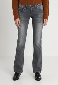 Jeans de mezclilla gris acampanados con bolsillos frontales, costuras en contraste y un ligero desgaste. Combinados con zapatos negros con acentos de tachuelas.