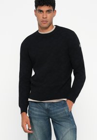 Ein junger Mann trägt einen schwarzen gemusterten Pullover über einem beigen Hemd und blauen Jeans. Er steht gegen einen weißen Hintergrund mit einer Hand in der Tasche.