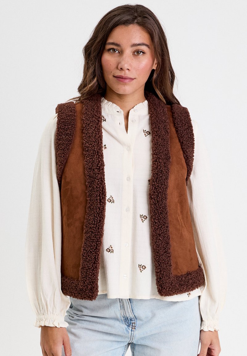 Gilet in finta suede marrone con un morbido bordo in shearling. Presenta un design semplice senza maniche, con una texture soffice e una vestibilità rilassata.