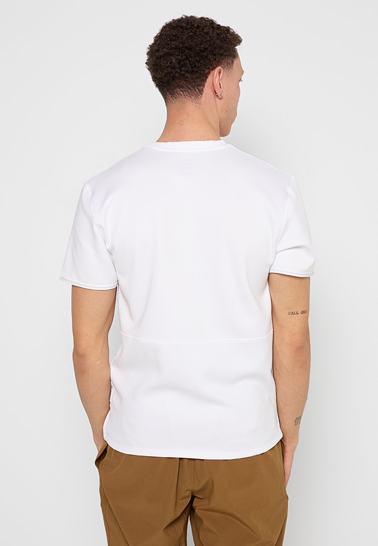 T-shirt blanc à manches courtes, présentant une texture lisse, un col rond et des détails de couture discrets à l'arrière. Associé à un pantalon marron.