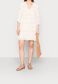Femme portant une robe florale légère et ample avec des plis, des sandales brunes à lanières, tenant un tote bag tissé beige à ses côtés.