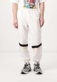 Pantalones joggers con cintura elástica blanca y franjas horizontales en negro y beige, con puños ajustados y una textura suave. Llevados con zapatillas grises y negras.