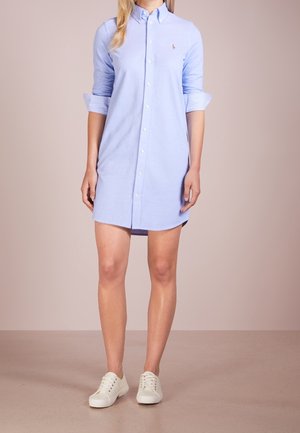 Nainen pukeutuneena vaaleansiniseen nappipaitamekkoon, jossa on käärityt hihat, ja valkoisiin tennareihin, neutraalia beigeä taustaa vasten.